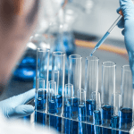 A close-up of a scientist in a lab using a pipette to add a blue liquid to a row of test tubes
